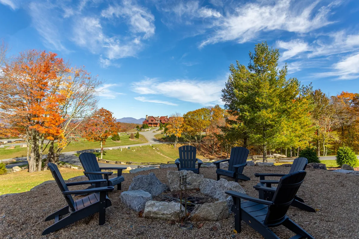 This exterior image features a cozy fire pit area surrounded by Adirondack chairs, situated under colorful autumn trees with distant mountain views. The open and inviting setup suggests a welcoming environment for outdoor relaxation and community gatherings.