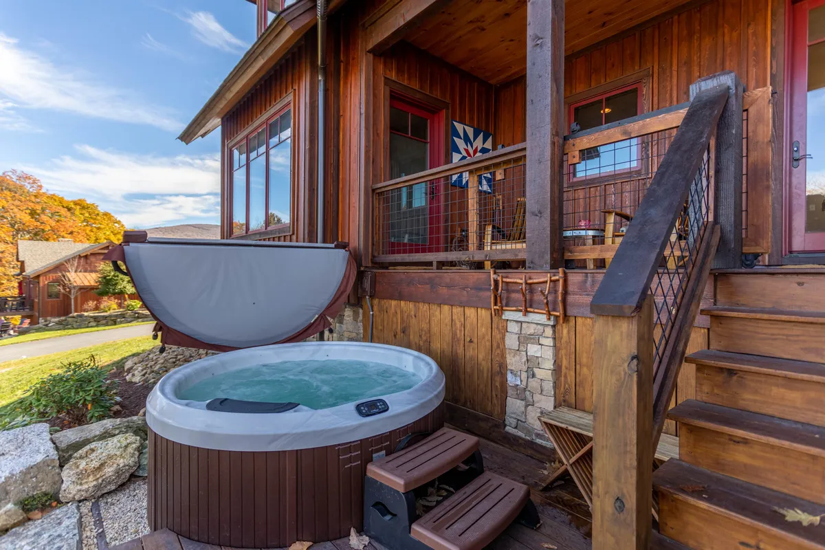 A cozy exterior shot of a bubbling hot tub on a wooden deck, with wooden steps and a decorative wall hanging visible in the background. The scene feels private and inviting, set against a tranquil outdoor backdrop.