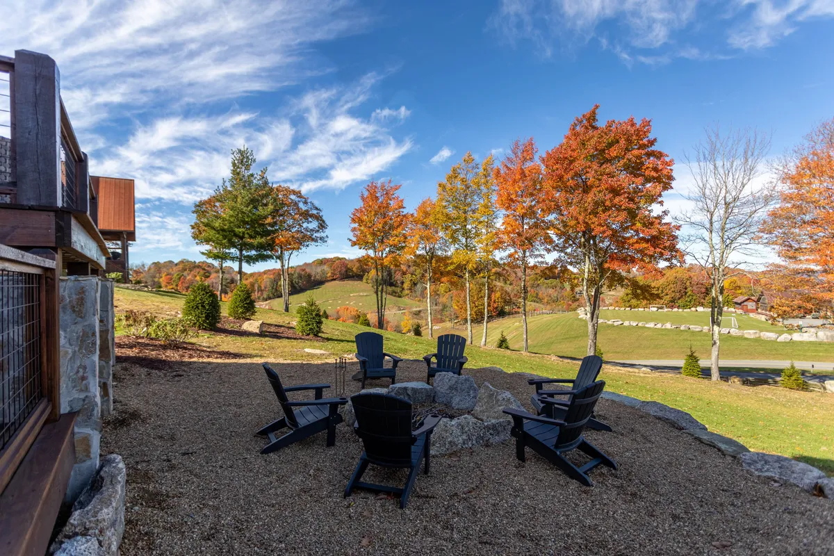 An inviting fire pit area surrounded by Adirondack chairs, framed by colorful autumn trees and rolling hills in the distance. The space is designed for gathering, storytelling, and enjoying the outdoors as a family.
