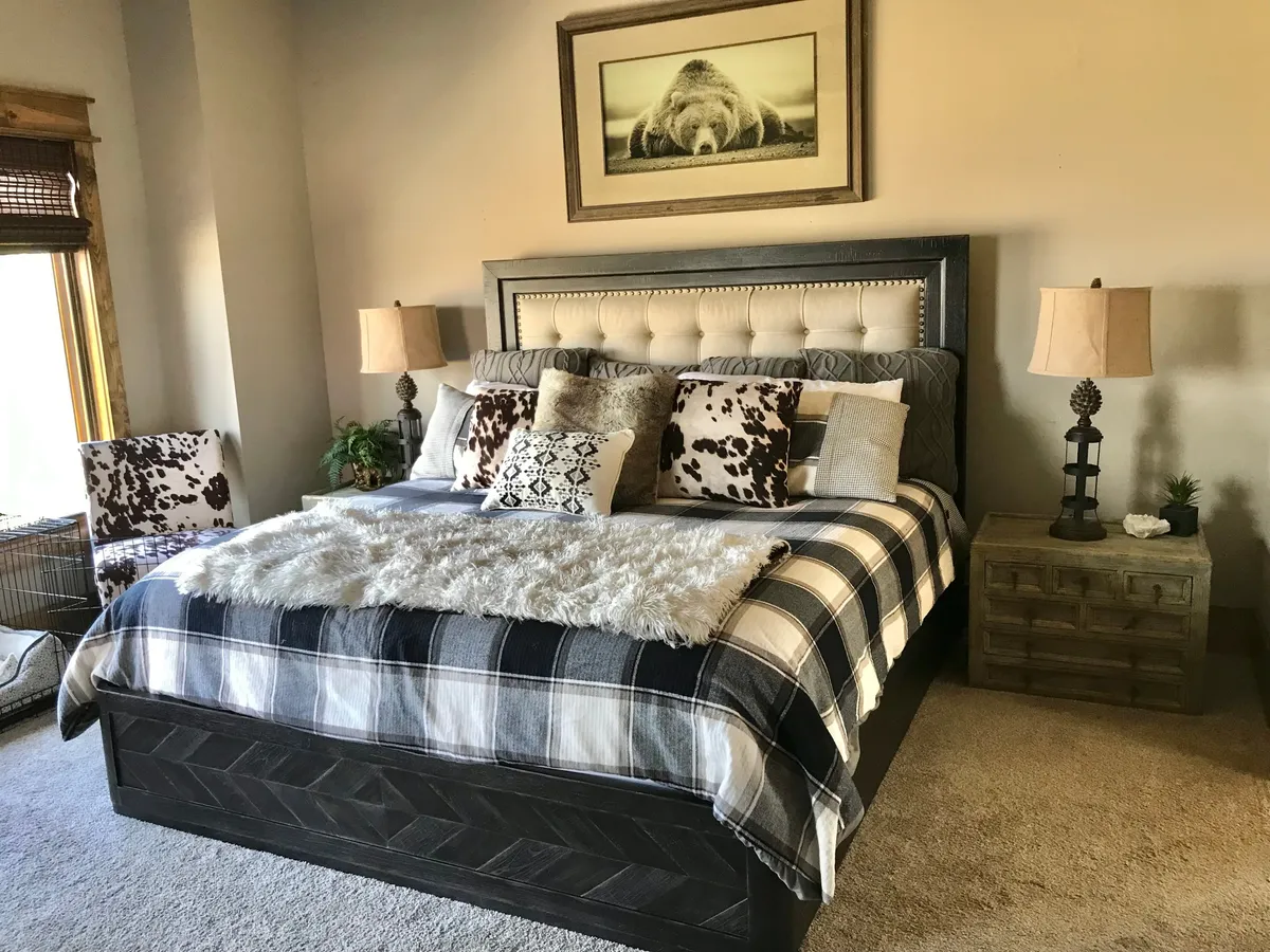 A spacious primary bedroom featuring a large bed with a tufted headboard, plush decorative pillows, a cowhide patterned chair, and framed bear art. The soft natural light streams through a nearby window, creating a tranquil and comfortable atmosphere ideal for relaxation.