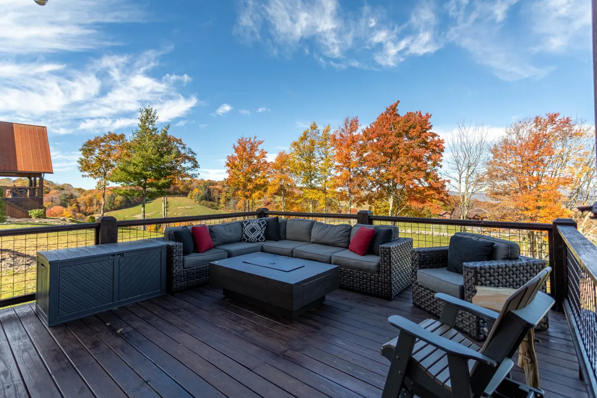 An outdoor seating area arranged around a fire pit, framed by autumn foliage and mountain views. Adirondack chairs invite guests to gather, while warm, golden light suggests the approach of sunset or evening.