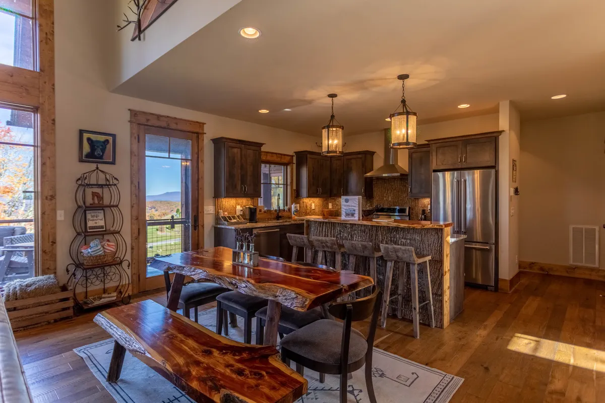 A view from the kitchen into the dining area reveals a large wooden dining table set near vast windows, capturing natural light and scenic outdoor views. The kitchen island with bar stools and pendant lighting connects the two spaces, showcasing the flow of the open-concept layout.