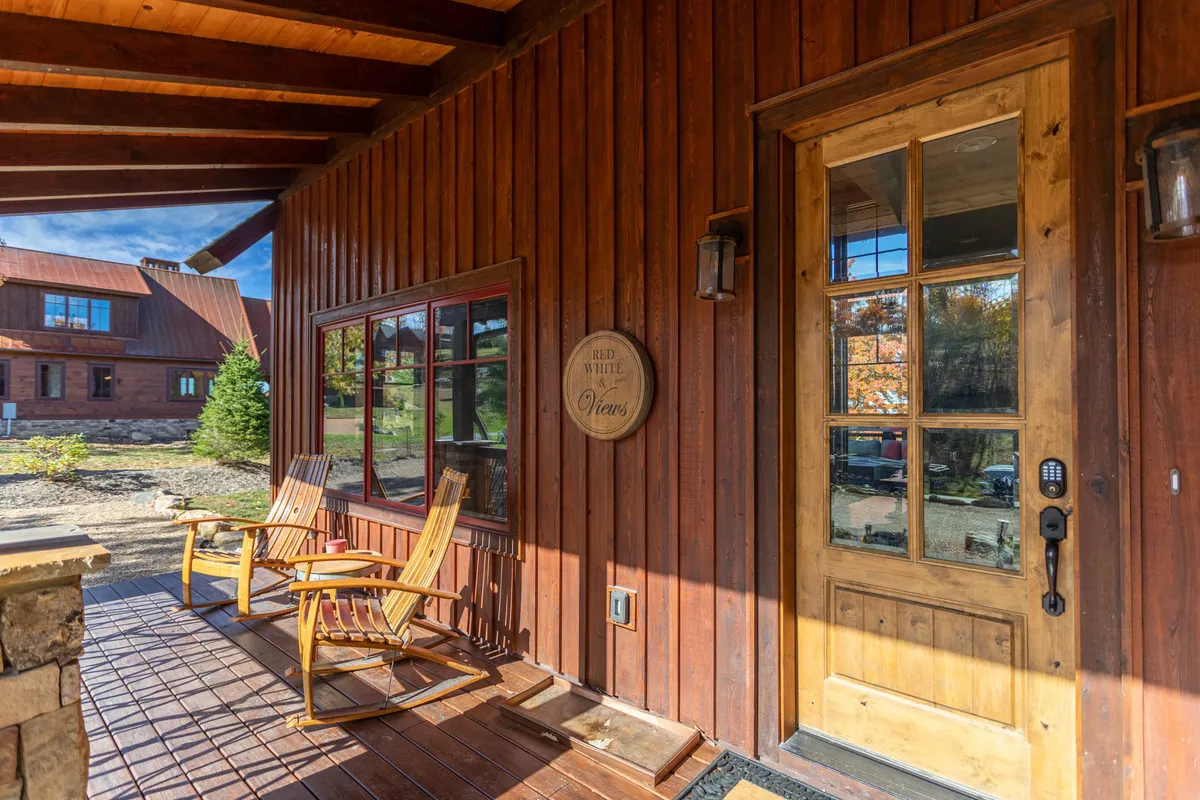 An exterior shot of the home's entrance, showcasing wooden siding, a welcoming wooden door with red trim, and cozy rocking chairs arranged on the porch. The setting suggests a pet- and family-friendly atmosphere and emphasizes the connection between indoor comfort and the surrounding landscape.