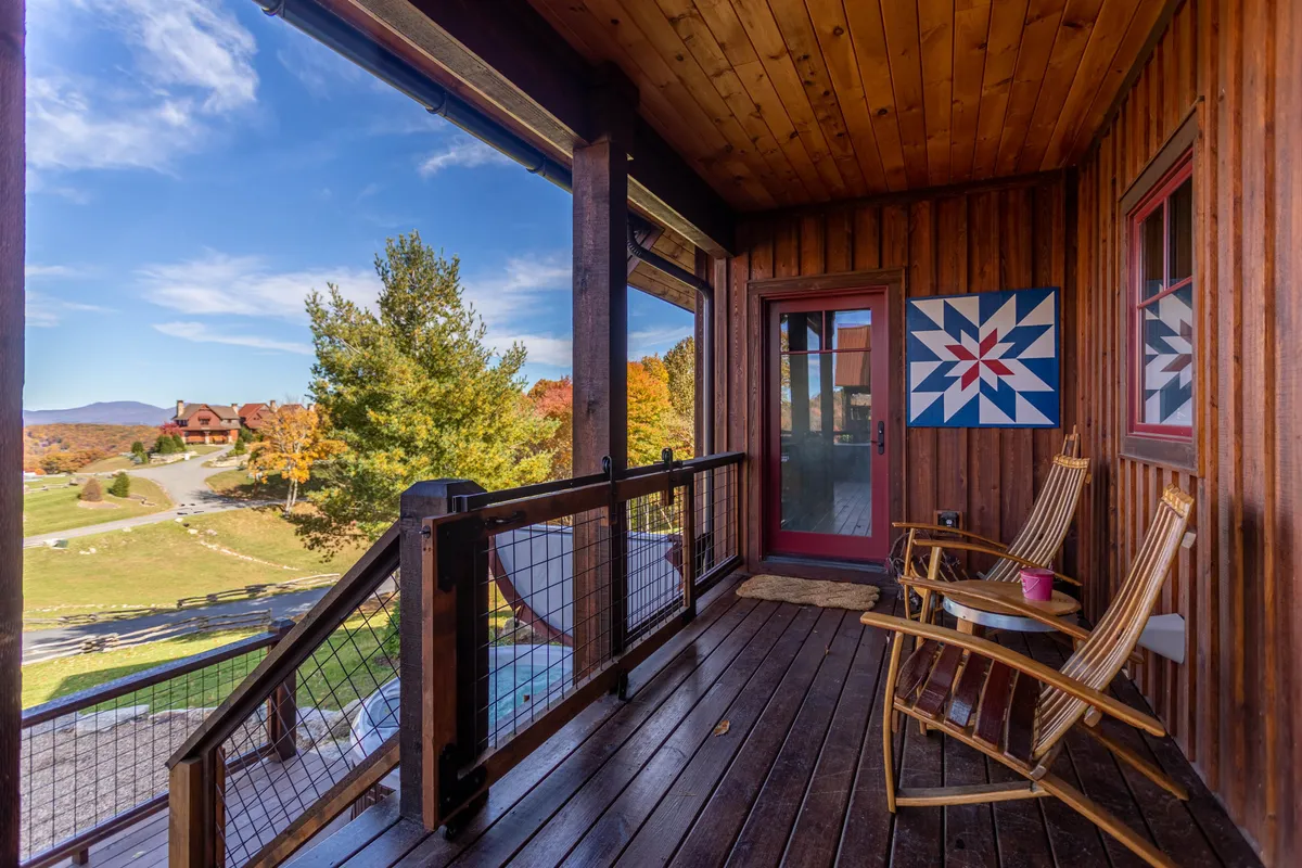 This exterior view centers on a porch with rocking chairs and quilt-pattern artwork, overlooking a scenic landscape. The relaxed setup evokes a sense of tranquility and enjoyment of the outdoors, ideal for unwinding with a pet after a walk.