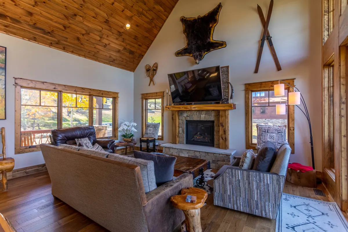 A grand living area featuring a rustic stone fireplace, plush sofas, exposed wooden beams, and expansive windows framing mountain views. A large TV is mounted above the mantel, creating a focal point for movie nights or gatherings. Warm wood accents and thoughtful decor enhance the inviting atmosphere.