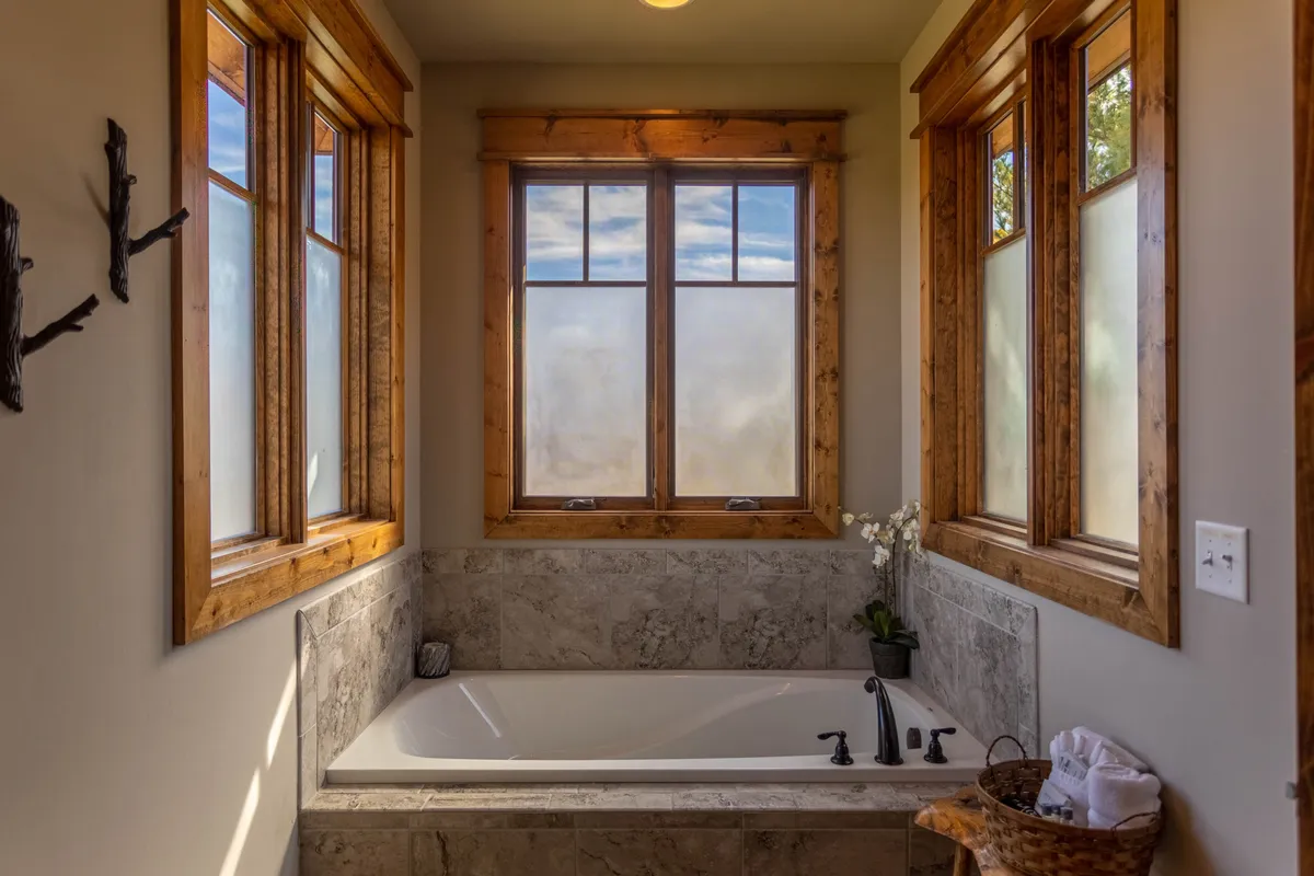 A serene bathroom featuring a standalone bathtub by a large window, wooden accents, and a floral arrangement. Natural light fills the space, drawing attention to the possibility of a relaxing bath with peaceful views of the outdoors.