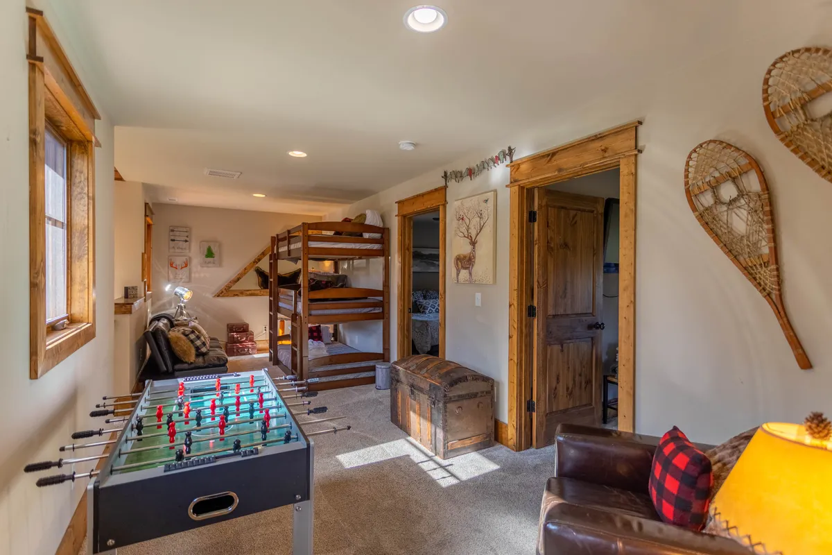 This image features a vibrant loft-like space with a foosball table as the centerpiece, flanked by cozy bunk beds, an armchair for reading or lounging, a rustic wooden chest, and decorative snowshoes on the wall. The ambient lighting and playful decor create a cheerful, inviting environment ideal for games and relaxation.