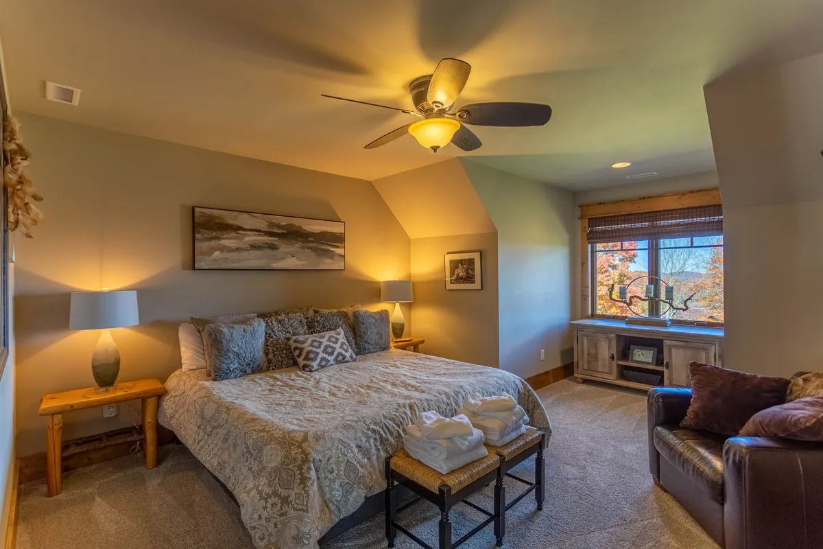 A restful bedroom with a comfortable bed adorned with decorative pillows, a window offering beautiful views, a side table with a lamp, and an armchair. The room exudes calm and is designed for unwinding, reading, or simply enjoying the scenery.