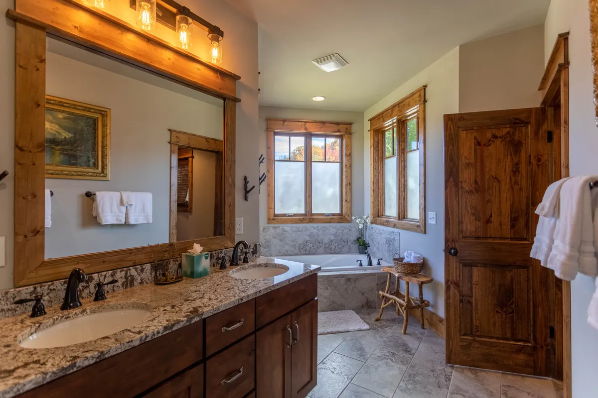 A serene bathroom with a double vanity and sinks, large mirrors, a soaking bathtub, and a window that lets in natural light. Wooden accents add warmth to the space, creating a spa-like environment perfect for unwinding after a long day.