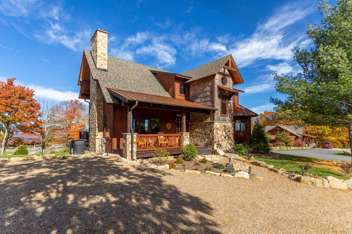 The image features a stunning stone and wood cabin perched amidst vibrant autumn foliage. The spacious wooden porch overlooks sweeping mountain views, inviting guests to gather outdoors, watch sunsets, and soak in the natural beauty. Large windows promise cozy interiors filled with light and connection to the scenic landscape.